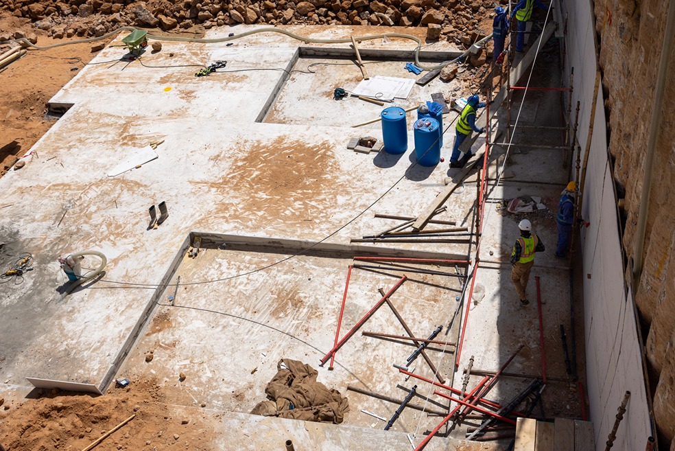 Cove Edition I By Imtiaz 1 Workers in safety gear are on a construction site, surrounded by tools, materials, and uneven terrain.